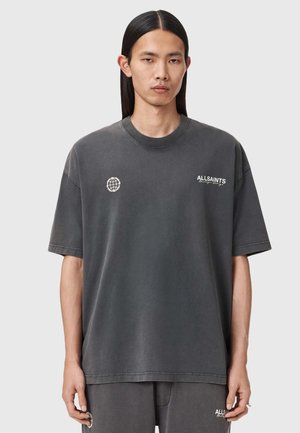 Ung person med langt sort hår iført en oversize mørkegrå AllSaints t-shirt og matchende shorts mod en ensfarvet baggrund.