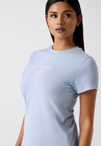 Jasnoniebieska koszulka z krótkim rękawem wykonana z miękkiego materiału, z białym haftowanym logo z przodu. Okrągły dekolt z minimalną ilością szwów.