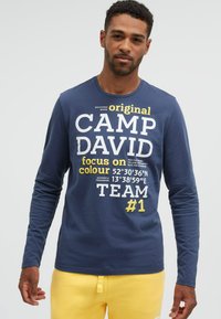 Mann trägt ein blaues Langarmshirt mit der Aufschrift "CAMP DAVID" und gelbem Text, kombiniert mit gelben Hosen vor einem schlichten hellen Hintergrund.