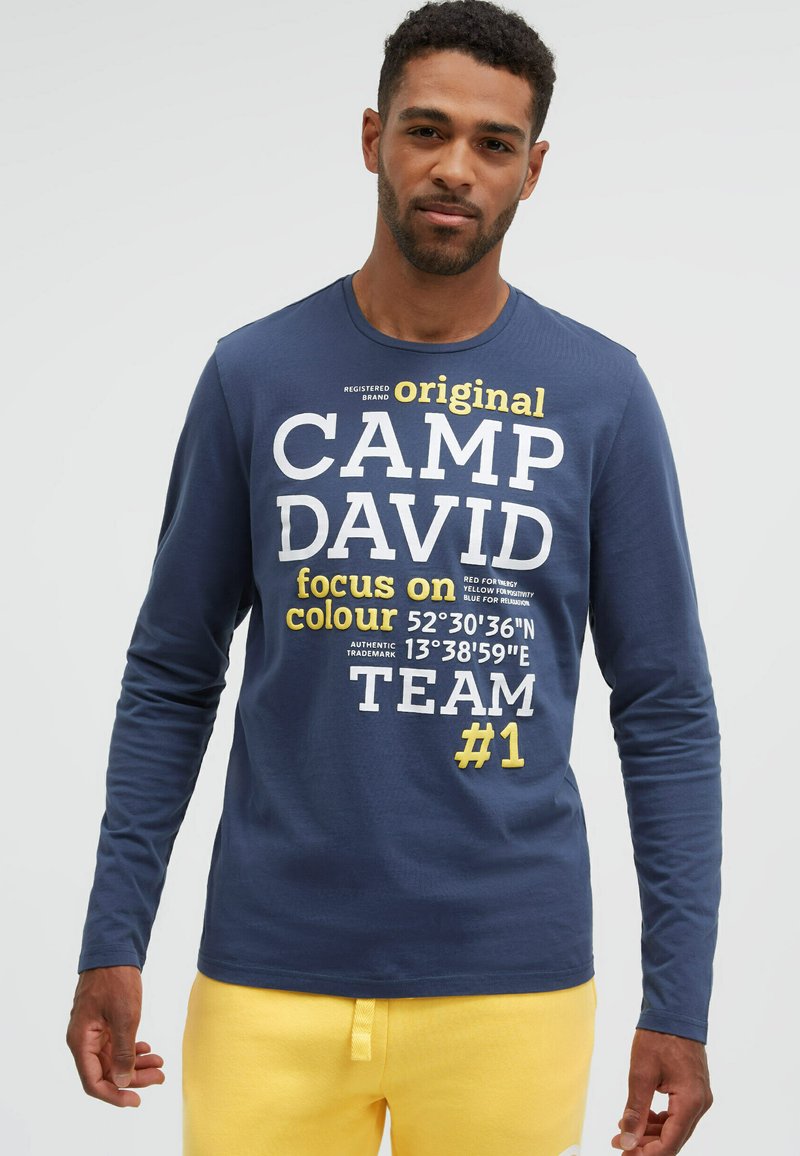 Mann trägt ein blaues Langarmshirt mit der Aufschrift "CAMP DAVID" und gelbem Text, kombiniert mit gelben Hosen vor einem schlichten hellen Hintergrund.