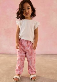 Haut blanc à manches courtes en côtes et pantalon cargo rose camouflage avec poches, associés à des sandales blanches sur un sol lisse avec un fond rose.