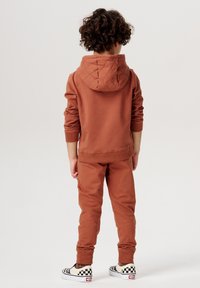 Roestkleurige trainingsoutfit met een gewatteerde hoodie, ribgebreide manchetten en bijpassende broek. Het kind draagt zwart-wit geblokte sneakers.