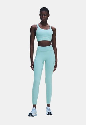 Frau steht und blickt nach vorne, trägt einen hellblauen Sport-BH, passende Leggings und weiße Sportschuhe vor einem schlichten weißen Hintergrund.