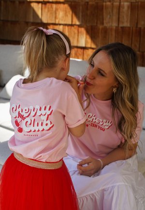 Zwei Personen tragen passende hellrosa T-Shirts mit der Aufschrift "Cherry Club". Eine trägt einen roten Tüllrock, die andere einen weißen Rock. Natürliche Umgebung.