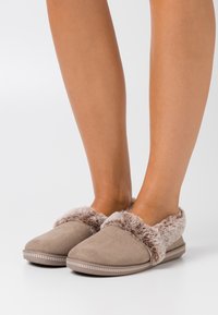 Bruine suede pantoffels met een pluizige bontafwerking en een ronde neus. De zool heeft een decoratieve stikseldetail. Over het geheel genomen een zachte textuur.