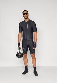 Černý cyklistický dres s krátkými rukávy a zipem vpředu, s bílým logem a akcenty. Model drží černou helmu a nosí sportovní sluneční brýle.