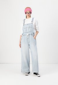 Salopette en denim bleu clair avec jambes larges, dotée d'une poche avant, de bretelles réglables et d'une taille avec cordon de serrage. Assortie à des baskets noires.