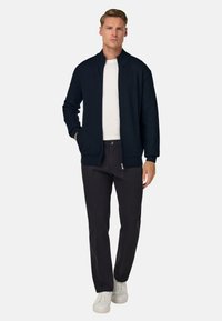 Temno modra rebrasta jakna z visokim ovratnikom, v kombinaciji s črnimi hlačami in belimi supergami, ustvarja plasten izgled v sproščenem slogu.