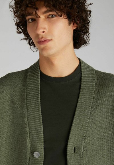 Cardigan lavorato a maglia di colore verde oliva con colletto a coste e chiusura con un bottone, indossato sopra una maglietta verde scuro, mostrando una texture liscia.