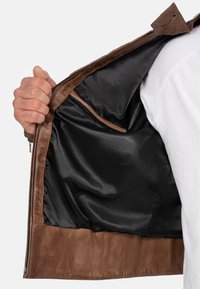 Interieur van een bruin leren jack, met een gladde zwarte voering en een bruin leren zoom. Ritsafsluiting met een subtiele zakdetail zichtbaar.