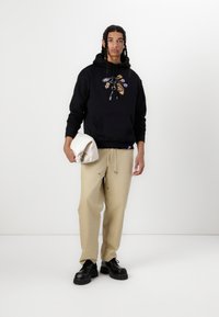 Schwarzer Hoodie mit Logo-Patches, Kängurutasche und Kordelzug. Beige Hose, lässiger Schnitt, und schwarze klobige Schuhe. Hält eine weiße Tasche.