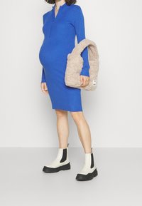 Blå ribbade stickade klänning med dragkedja vid kragen, kombinerad med en fluffig beige axelväska och vita anklarstövlar med svarta sidopaneler.