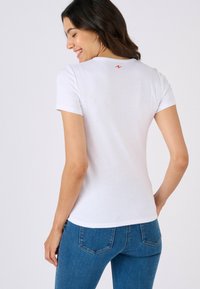 T-shirt blanc à manches courtes en tissu doux, avec un petit logo rouge au dos. Associé à un jean bleu moulant.
