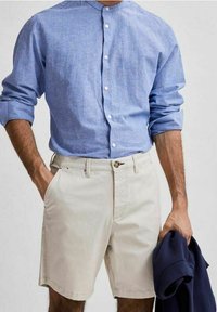 Camicia azzurra a righe con maniche arrotolate, abbinata a pantaloni corti bianchi. La camicia ha un collo rotondo e l'outfit include un blazer blu navy.