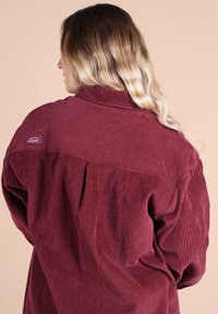 Chemise en velours côtelé bordeaux avec un col pointu, tissu texturé et une petite étiquette logo sur l'épaule ; présente un détail de plis à l'arrière.