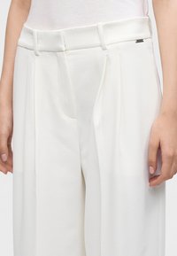 Gros plan sur un pantalon blanc ajusté avec passants de ceinture et plis latéraux, porté avec un haut blanc, montrant les mains reposant sur les côtés.