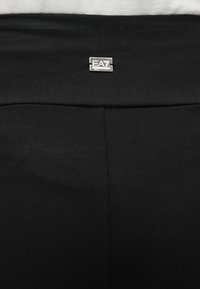 Pantaloni atletici neri con una texture liscia. Presentano un'etichetta con il logo argentato EA7 sulla vita. Design semplice, senza motivi o accenti aggiuntivi.