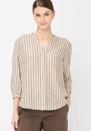 Femme portant un chemisier beige à rayures blanches verticales avec manches trois-quarts et un pantalon marron, debout devant un fond blanc.