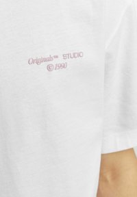 Camiseta de algodón blanca con el texto bordado en rosa "Originals��™ STUDIO ©1990" en la parte frontal. Diseño de mangas cortas y cuello redondo.