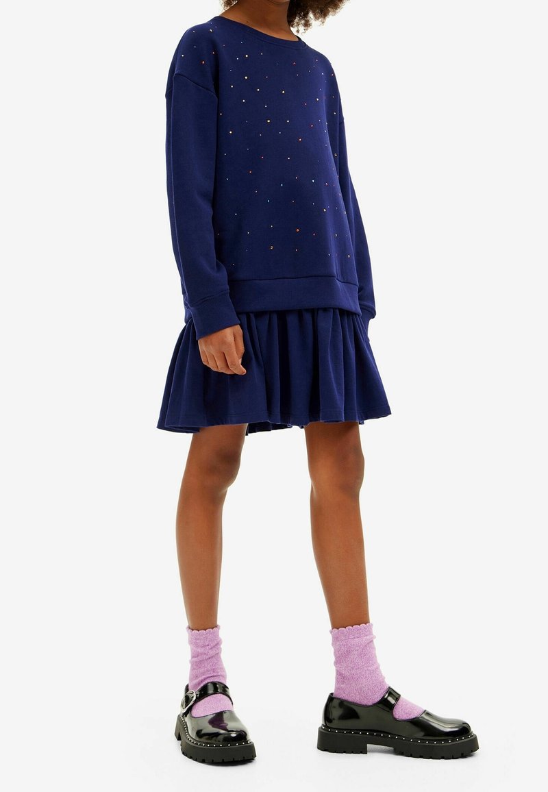 Personne portant un pull bleu marine à petits points colorés, une jupe plissée assortie, des chaussettes roses et des chaussures Mary Jane noires.