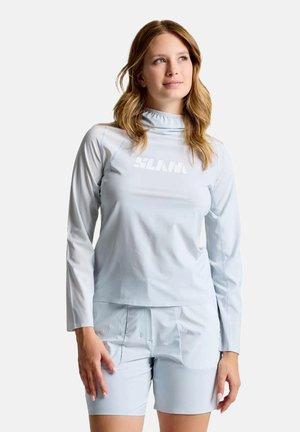 Woman with wavy hair wearing a light gray long-sleeve top and matching shorts, looking slightly upward and to the side.