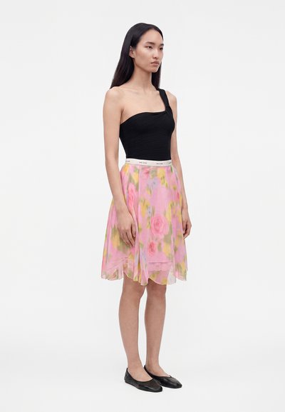 Femme debout en haut noir asymétrique, jupe rose fleurie longueur genou avec ceinture blanche et ballerines noires, sur un fond uni.
