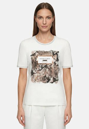 Femme portant une chemise blanche à manches courtes avec un motif abstrait marron et noir et le texte « GOOD VIBES » sur le devant, associée à un pantalon blanc.