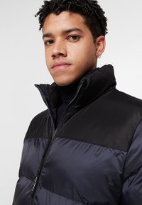 Gevoerde jas met een zwart bovenstuk en een donkergrijs onderstuk, voorzien van een hoge kraag, een ritssluiting aan de voorkant en een geribbeld patroon.