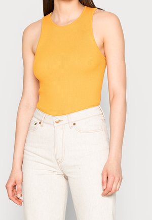 Mujer con una camiseta naranja sin mangas acanalada metida en unos vaqueros beige claro de talle alto, de pie con los brazos relajados a los lados.