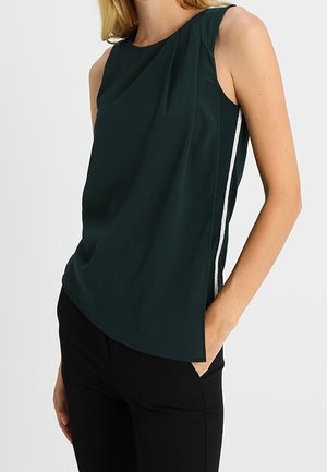 Blouse sans manches vert foncé avec un détail de pli sur le côté et une rayure claire, portée avec un pantalon noir, la main dans la poche.