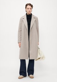 Kvinne som står iført en beige lang frakk, svart genser med høy hals, vide jeans, beige sneakers, holder en hvit posete håndveske.