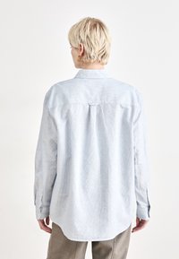 Camicia azzurra chiaro con bottoni in un tessuto leggero e testurizzato, caratterizzata da un orlo curvo e maniche lunghe con polsini a bottone.