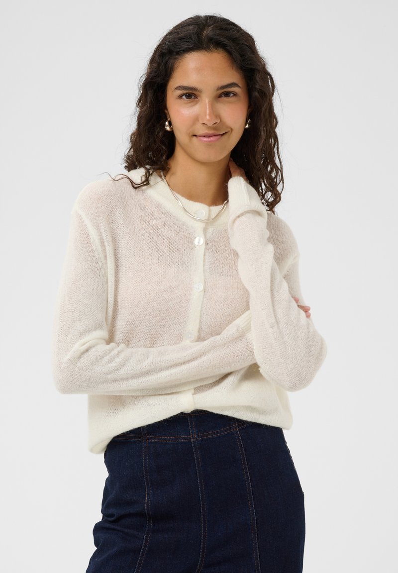 Femme aux longs cheveux bouclés portant un cardigan crème et un jean foncé, souriant doucement avec une main touchant son cou sur un fond uni.
