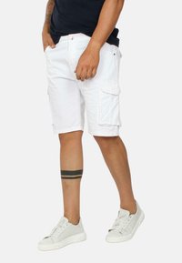 Pantaloni cargo bianchi con tasche laterali e chiusura a pulsante, abbinati a sneakers grigie. Il tessuto appare leggero e informale.