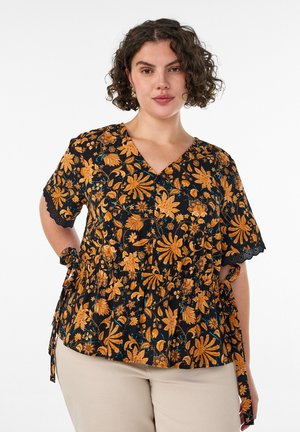Femme aux cheveux bouclés et foncés portant un chemisier noir avec des motifs floraux orange et un pantalon beige, debout devant un fond blanc uni.