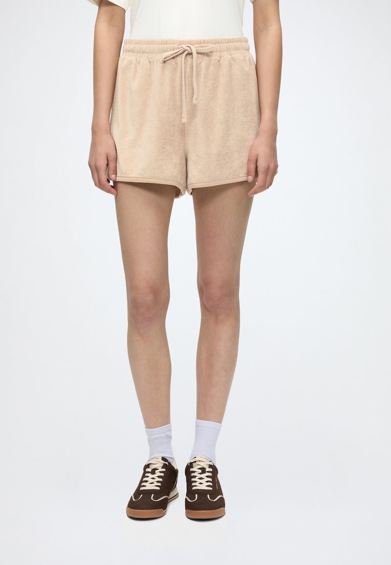 Personne portant un short beige à cordon, des chaussettes blanches et des baskets marron, debout devant un fond uni.