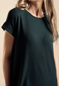 Tummanvihreä, ribballinen pusero, jossa on lyhyet käärityt hihat ja pyöreä kaula-aukko. Pehmeä tekstuuri, rento istuvuus, vähäinen yksityiskohtaisuus ja sileä laskos.