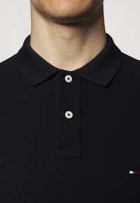 Granatowa koszulka polo z teksturowanej tkaniny z kołnierzykiem, wyposażona w dwa kremowe guziki oraz małe logo w kolorach czerwonym, białym i niebieskim.