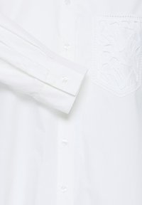 Chemise blanche boutonnée à manches longues avec une poche brodée texturée sur la poitrine gauche.