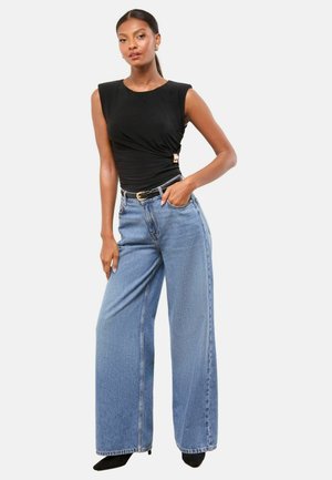 Schwarzes, eng anliegendes, ärmelloses Oberteil mit Wickeldetail und goldenen Akzenten, kombiniert mit hoch taillierten, weit geschnittenen hellblauen Jeans und schwarzen spitzen Absätzen.