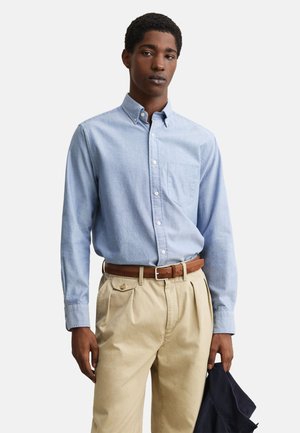 Jonge man die een lichtblauw overhemd met knopen en beige broek met een bruine riem draagt, houdt een donker marineblauw jasje in zijn linkerhand.