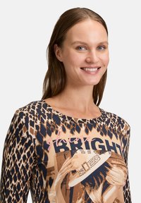 Długi rękaw bluzki z nadrukiem w panterkę w odcieniach brązu i granatu, z graficznym wzorem oraz różowym napisem "świeć JASNO".