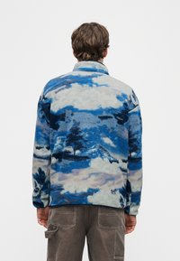 Άτομο που φοράει ένα μπλε και γκρι φλις μπουφάν με μοτίβο τοπίου με σύννεφα και δέντρα, συνδυασμένο με γκρι παντελόνι cargo, κοιτάζοντας προς τα πίσω.