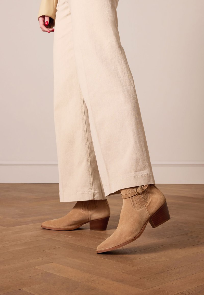 Stivaletti in suede beige con punta affusolata, tacco basso e spesso, e dettaglio a coste fissato con una fibbia sottile. Pantaloni a gamba larga color crema.