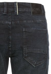 Ciemnoniebieskie dżinsy z fakturowanym wykończeniem, kieszeń naszywana o kanciastym kształcie oraz skórzana metka na pasku.