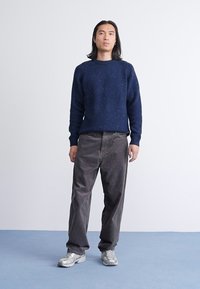 Marineblauwe gebreide trui met ribbeltextuur, gecombineerd met grijze ribfluwelen broek. Het model draagt zilverkleurige sportschoenen tegen een neutrale achtergrond.