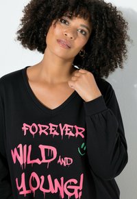 Maglietta nera a maniche lunghe con scollatura a V, con un audace testo in stile graffiti rosa "FOREVER WILD AND YOUNG!" e un piccolo dettaglio di faccina sorridente verde.
