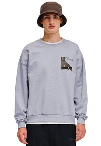 Grå sweatshirt med en avslappnad passform, ribbade muddar och midja. Har ett märke med svarta och gula grafiker på bröstet.