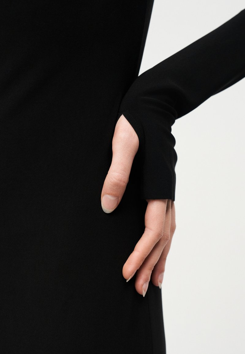 Main droite insérée dans l'ouverture du pouce d'une longue manche noire, sur fond blanc.