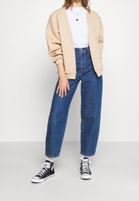 Cárdigan beige extragrande, camiseta blanca, jeans rectos de tiro alto azules y zapatillas negras altas con suelas y cordones blancos.